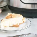 Instant Pot Traditional Cheesecake sliced and served on a white dessert plate sitting next to a silver fork in front of the Instant Pot