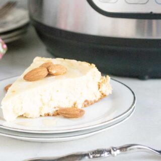 Instant Pot Traditional Cheesecake sliced and served on a white dessert plate sitting next to a silver fork in front of the Instant Pot