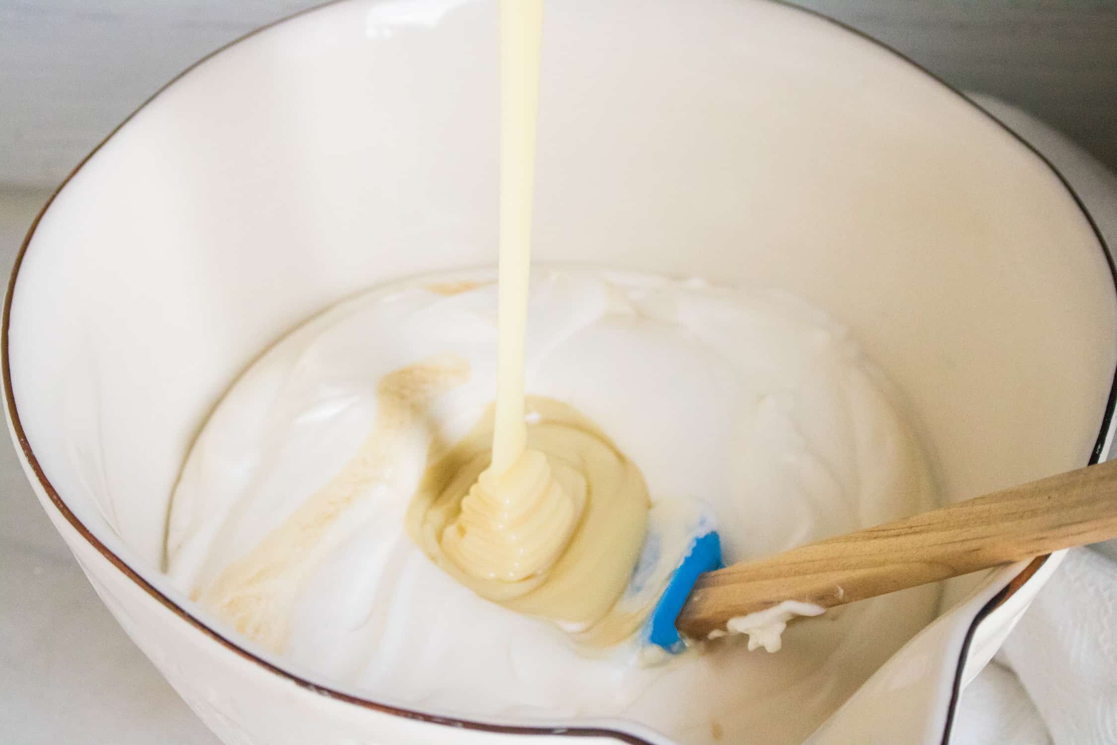 sweetened condensed milk being poured into white bowl filled with whipped heavy whipping cream for raspberry chocolate chunk no-churn ice cream