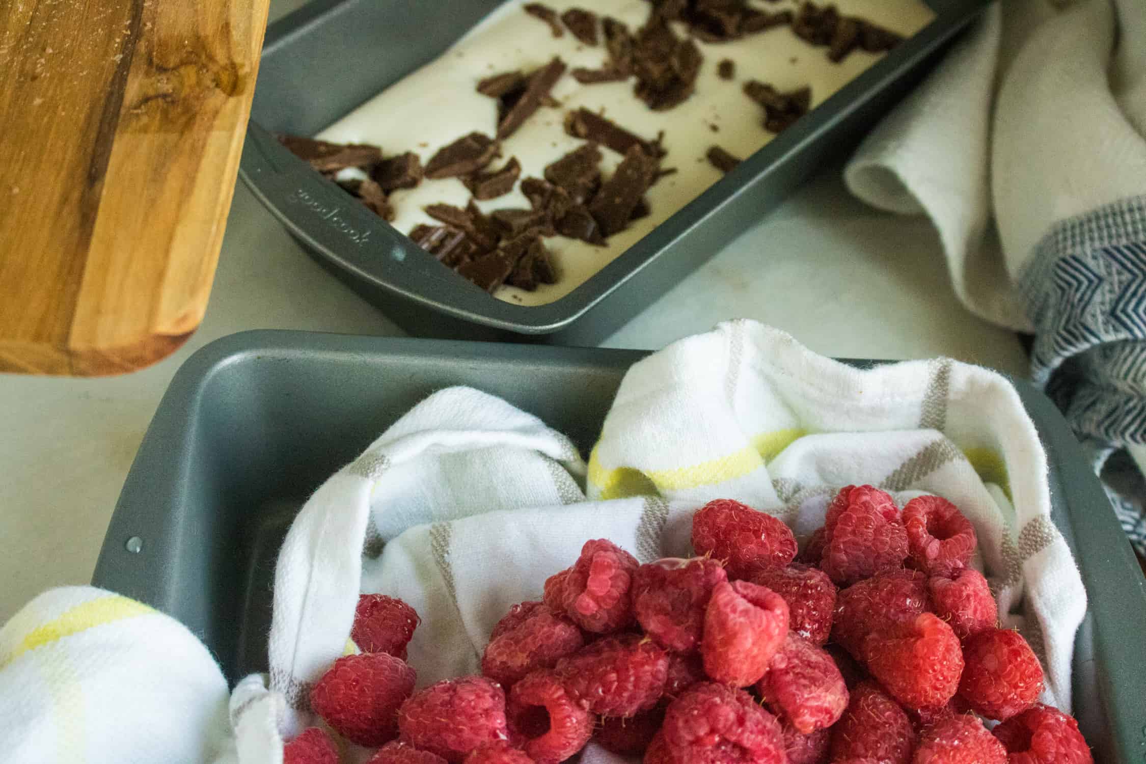 silver pan with white dishtowel liner filled with fresh raspberries and silver pan in background with whipped cream and chocolate chunks for raspberry chocolate chunk no-churn ice cream