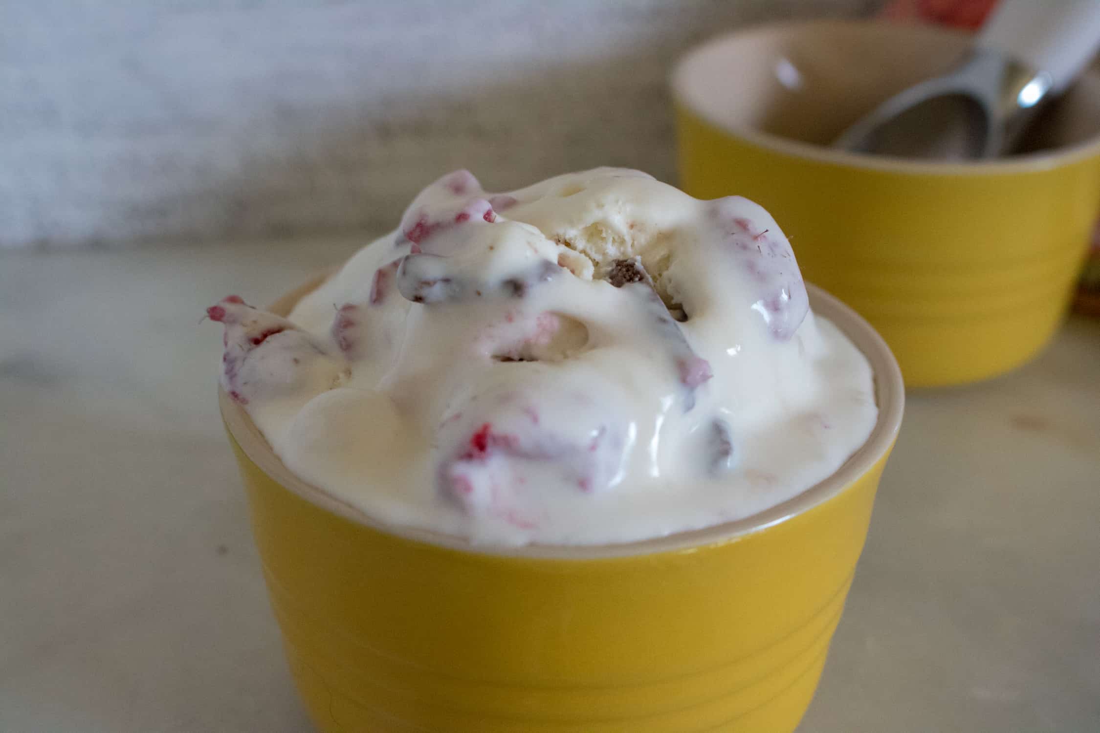 one scoop of Raspberry Chocolate Chunk No-Churn Ice Cream served in a yellow bowl with another yellow bowl in background with silver scoop in it