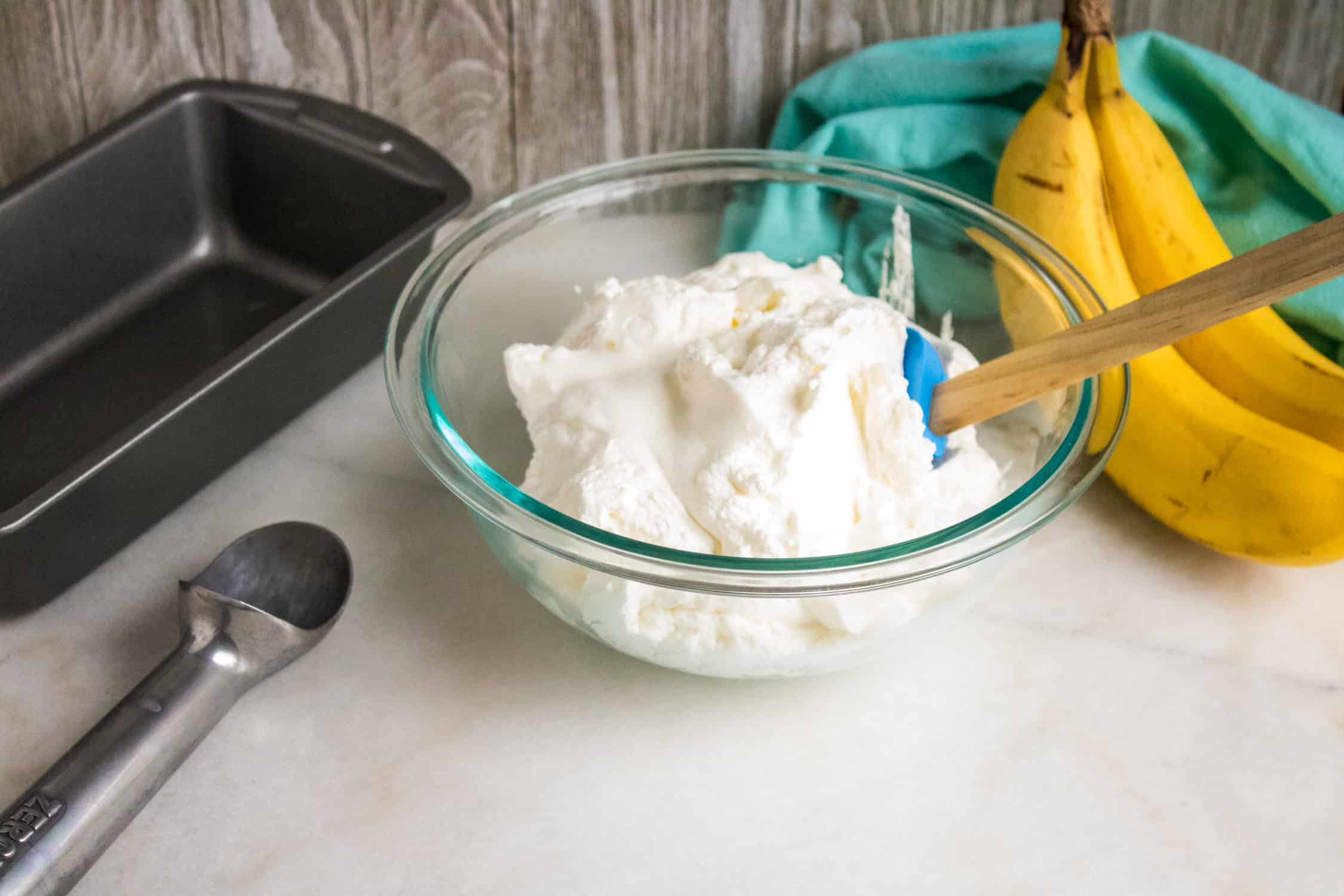 thick, heavy whipping cream in a glass bowl set next to two bananas, blue cloth napkin, freezer safe container and an ice cream scoop