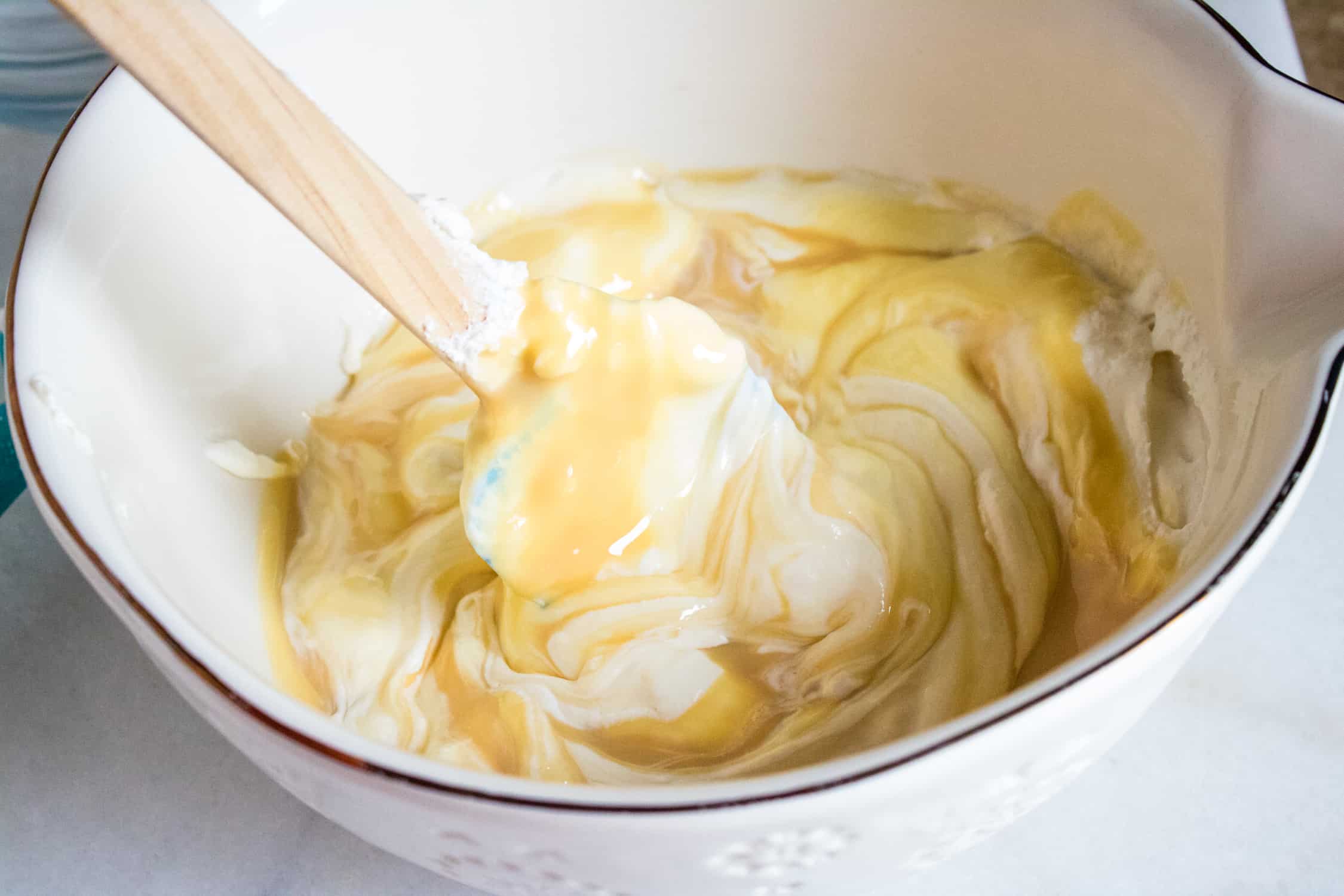 Whipped cream and sweetened condensed milk being mixed together in a white bowl with a spatula