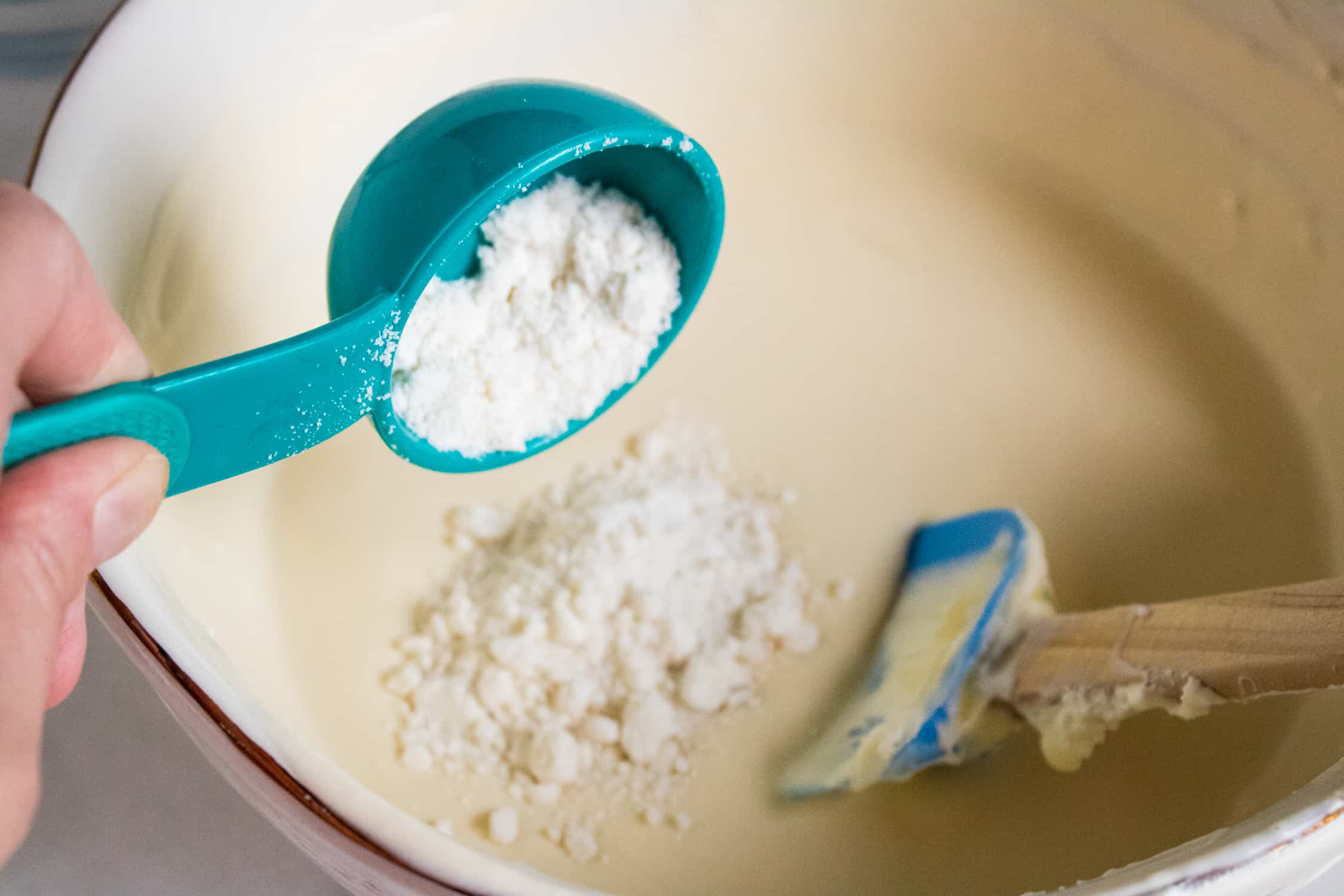 Birthday cake flavoring in a blue measuring spoon being added to the ice cream base in a bowl