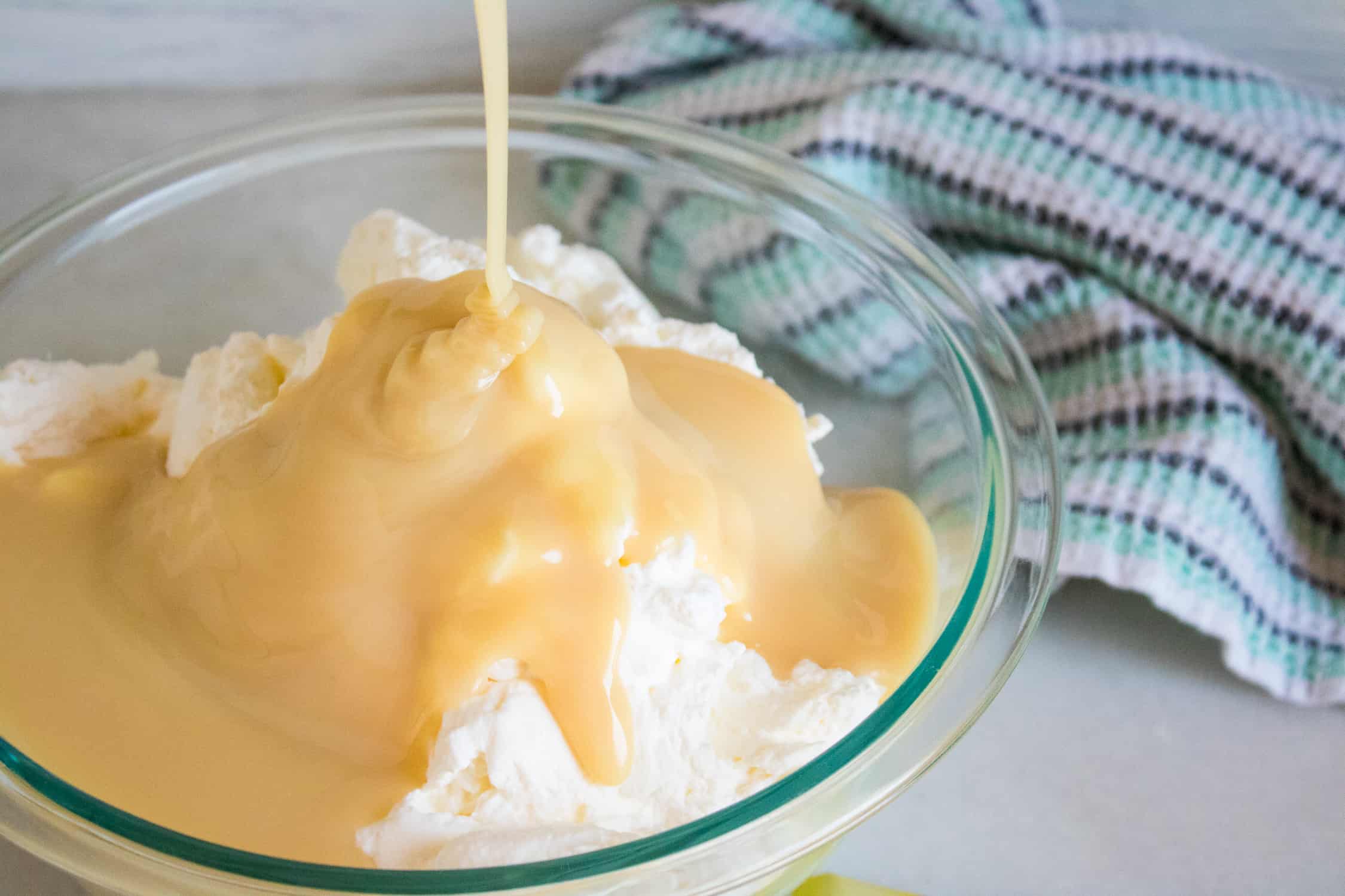 condensed milk being poured on top of the whipped cream inside a clear glass mixing bowl