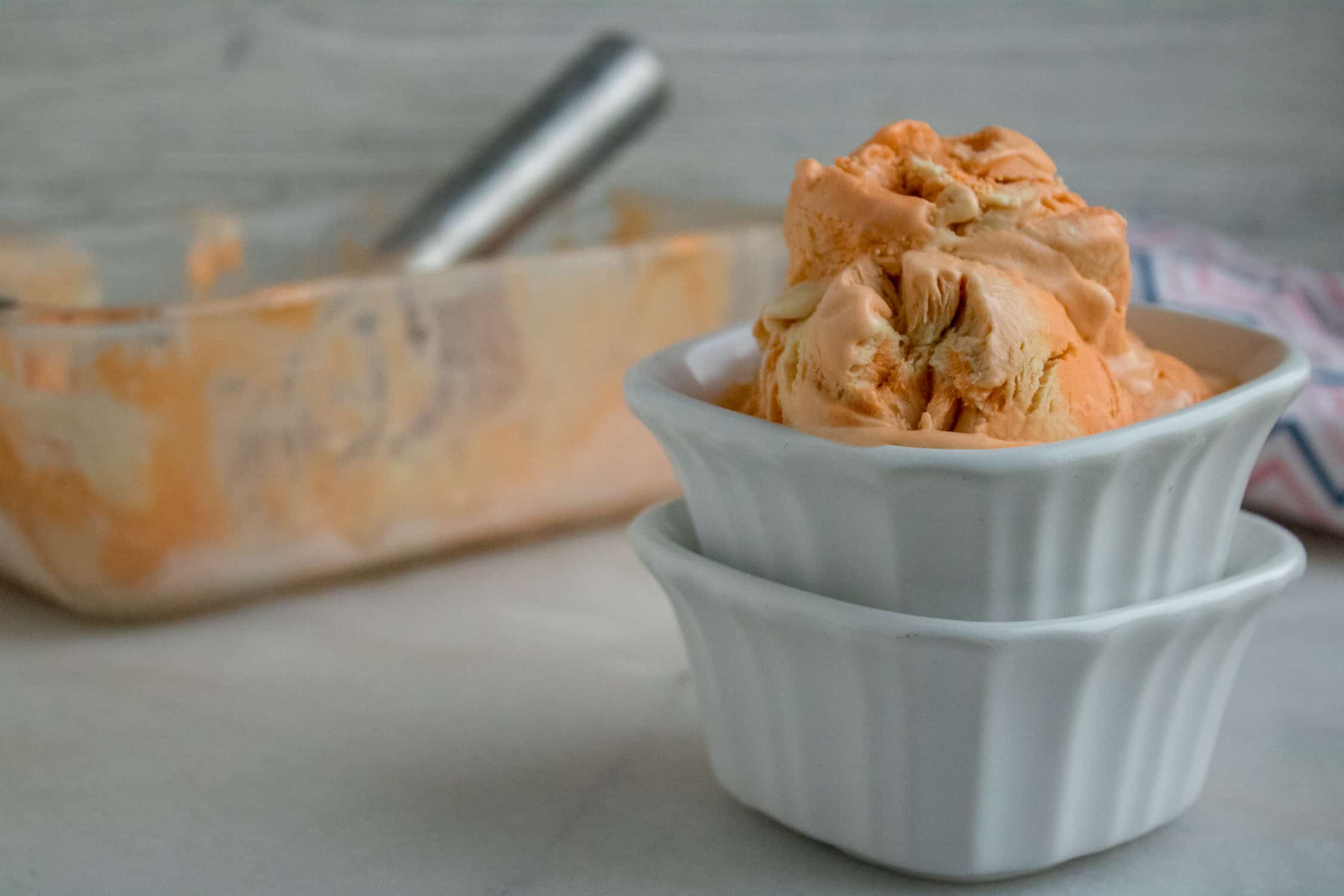Glass container of Orange Creamsicle No-Churn Ice Cream in front of a white square ceramic ramekin filled with two scoops of the ice cream