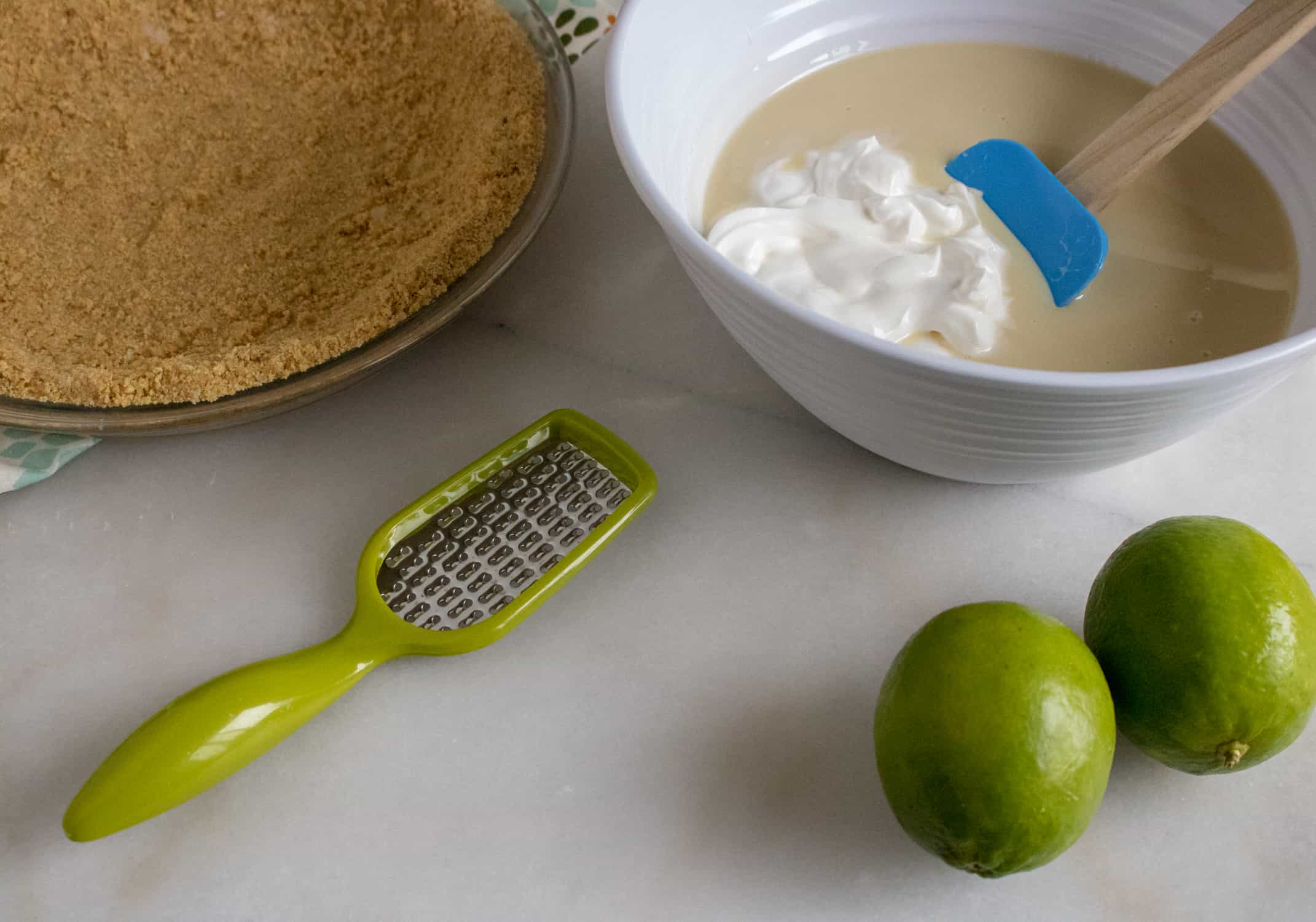 Graham cracker pie crust beside a white bowl with sweetened condensed milk and sour cream. Lime zester and two limes beside the bowl