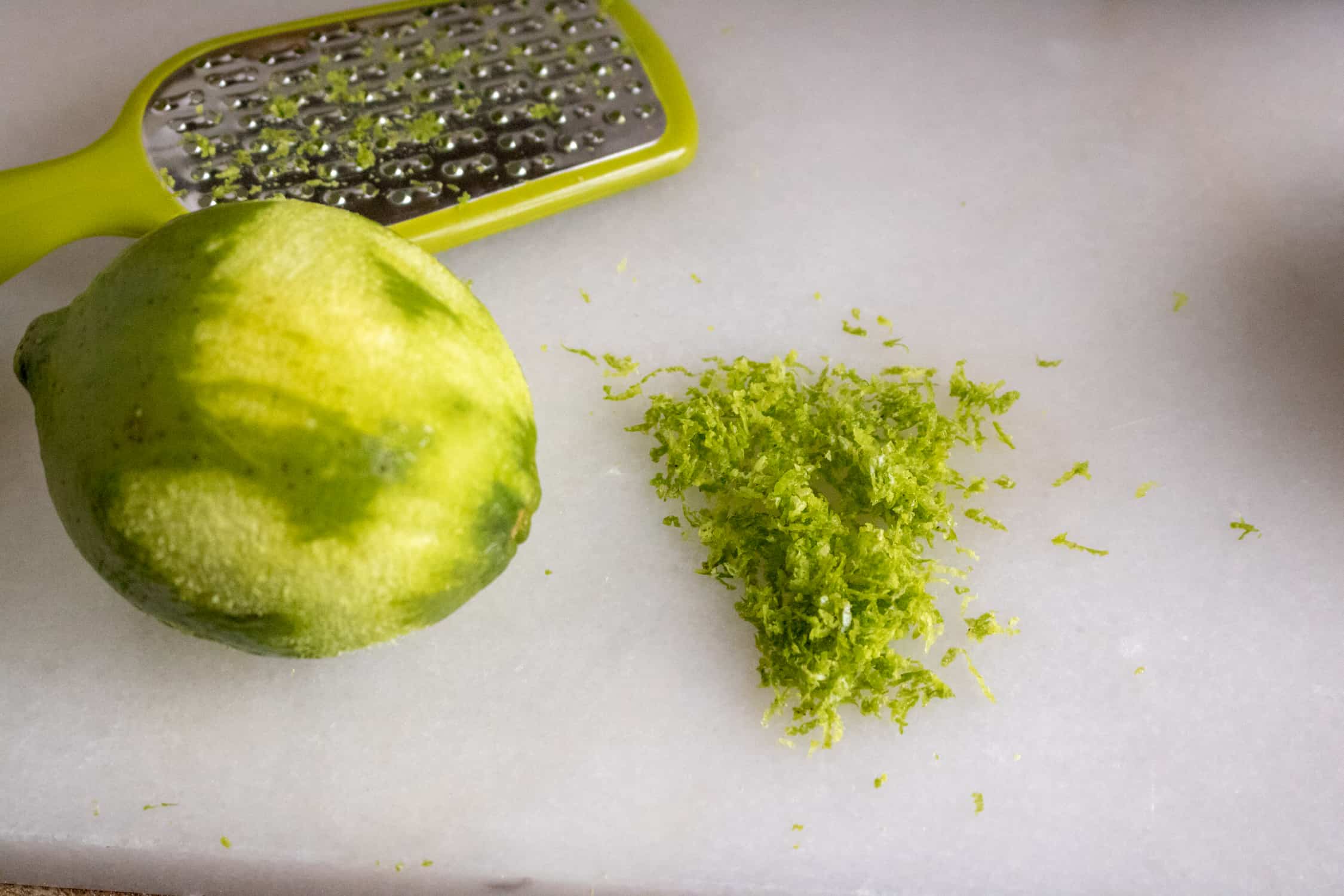 Zest grater beside a lime with zest removed and a pike of lime zest on a work surface