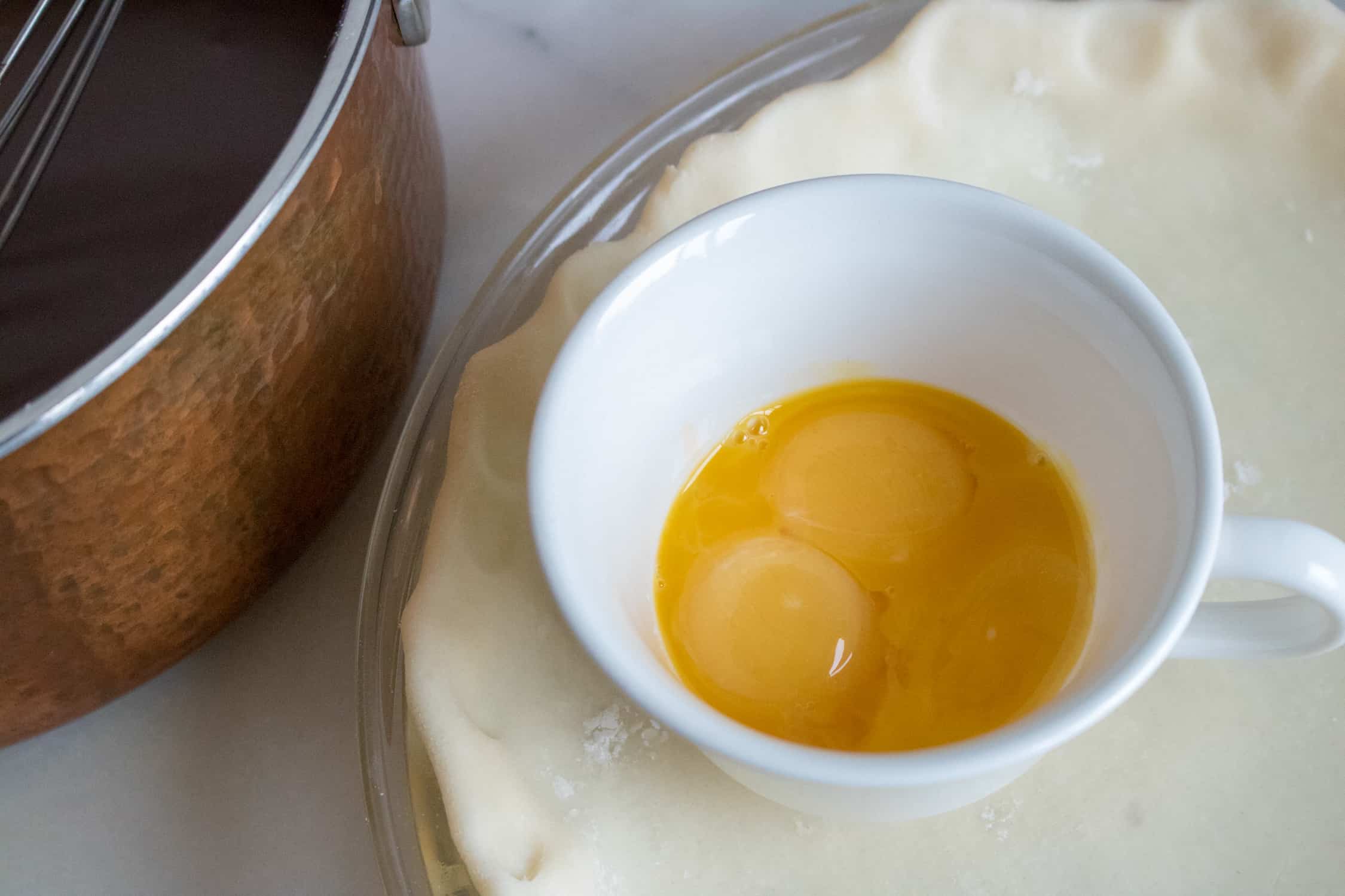 Three egg yolks in a white mug sitting on top of an unbaked pie shell with a sauce pan of chocolate pie filling to the left
