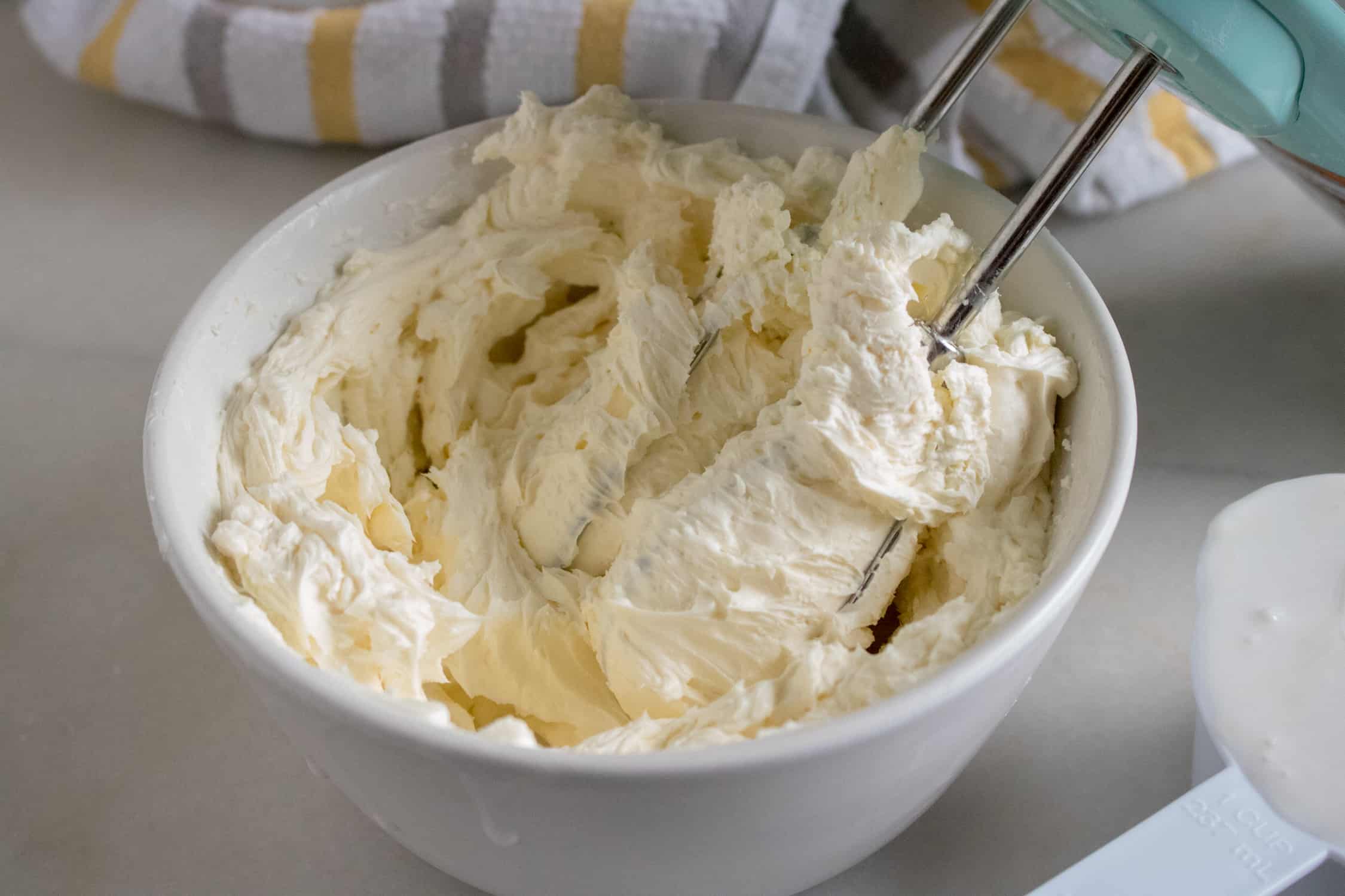 White bowl of cream cheese whipped until fluffy with electric mixer and striped towel in background