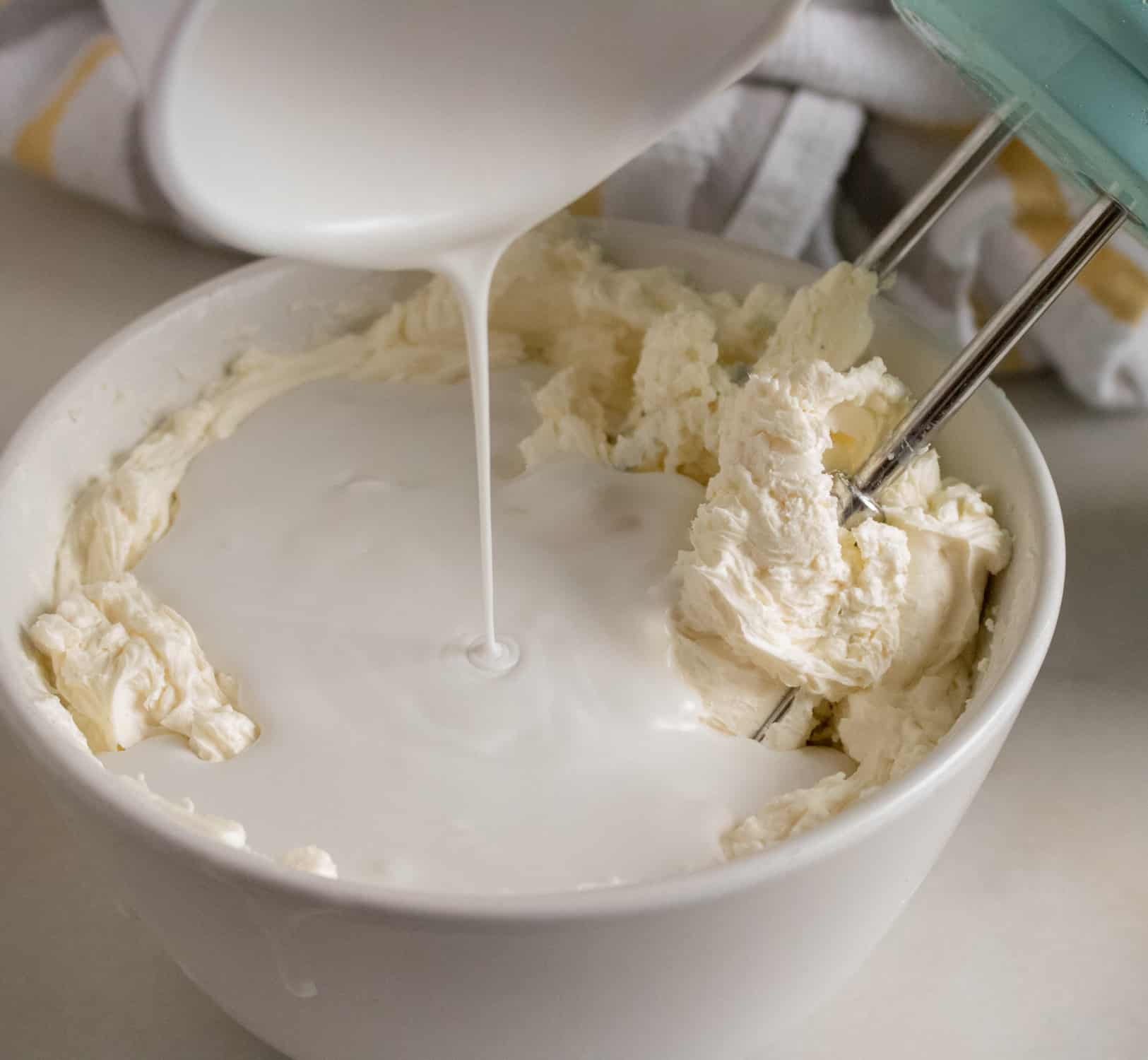 Coconut cream being added to whipped cream cheese in a white bowl with an electric mixer and striped towel in background