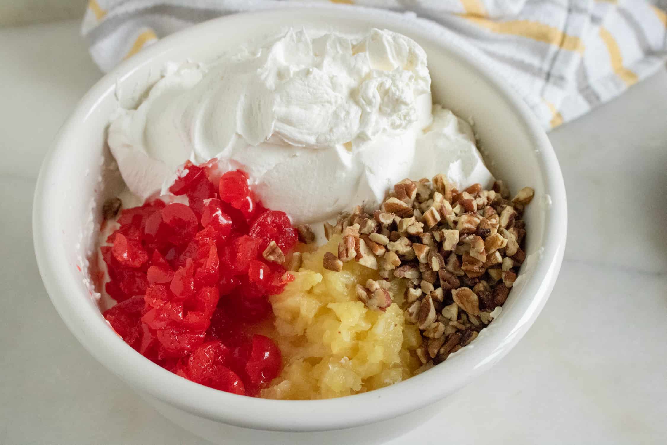 White bowl filled with whipped topping, chopped cherries, crushed coconut and chopped pecans