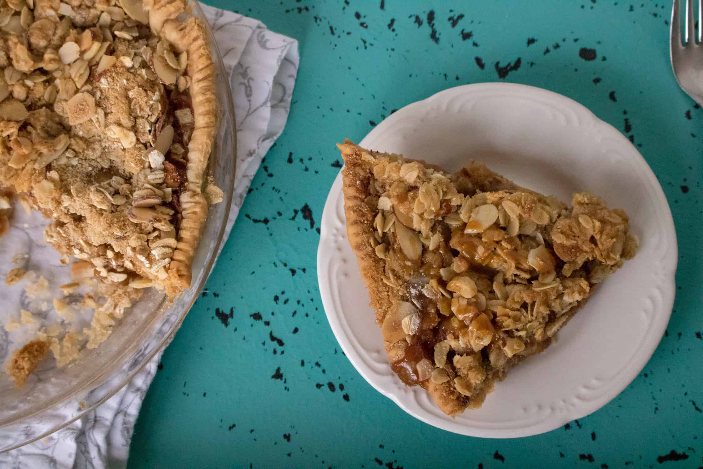 One slice of Caramel Apple Crumble Pie on a white plate with baked pie to the left. Both are sitting on a blue and black placemat