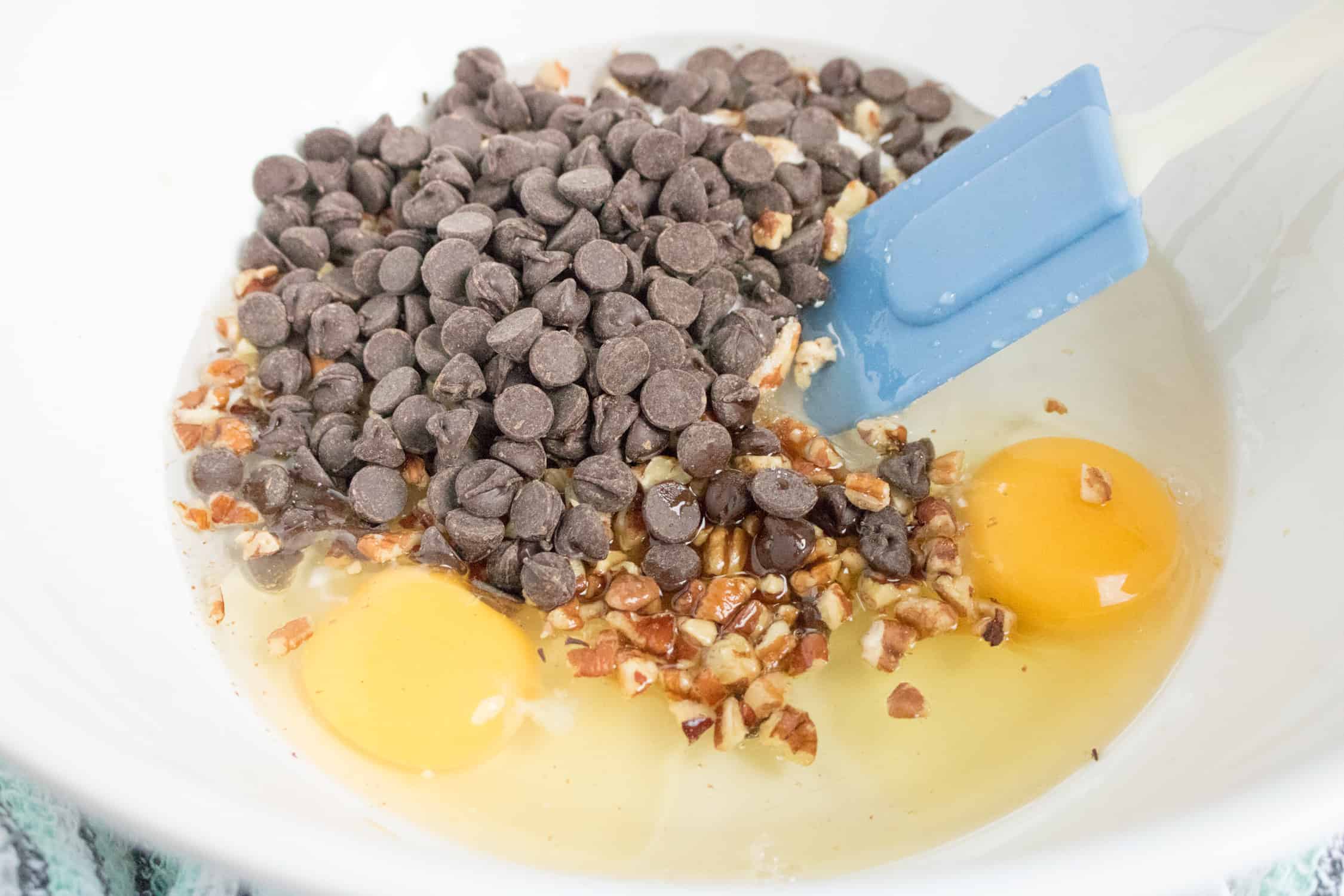 Looking down into a white bowl with chocolate chips and eggs for topping for Instant Pot Pecan Cheesecake with Chocolate Chips being mixed with a blue spatula.
