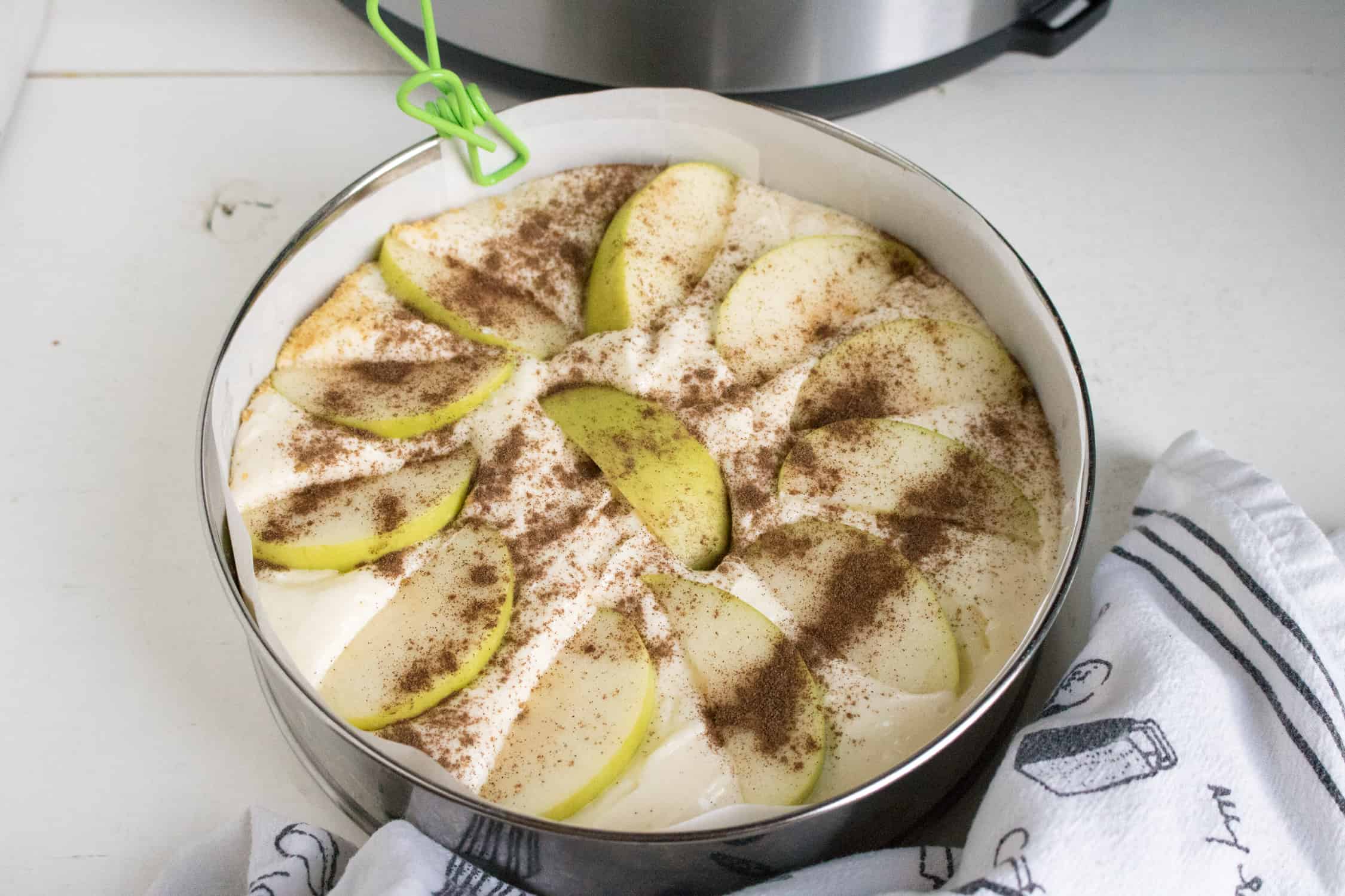  Instant Pot Apple Cheesecake in instant pot spring form pan with sliced apples on top, instant pot in back and black and white dishtowel in front