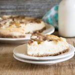 One slice of Instant Pot Apple Cheesecake cut from the whole cheesecake and served on a white dessert plate with a silver fork, glass of milk and blue print cloth napkin