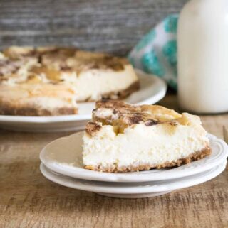 One slice of Instant Pot Apple Cheesecake cut from the whole cheesecake and served on a white dessert plate with a silver fork, glass of milk and blue print cloth napkin