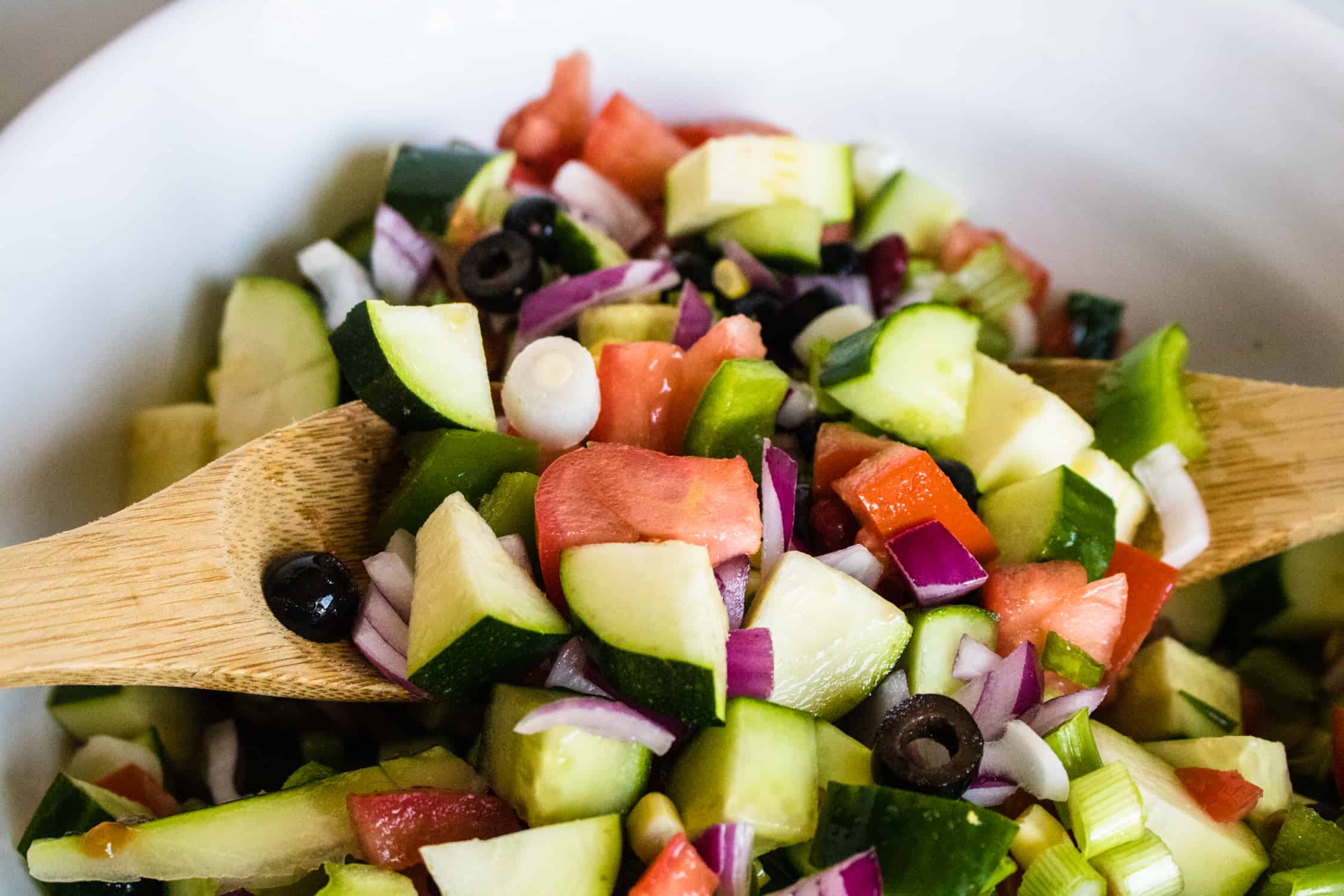 Zucchini, tomatoes, red and green onions, beans and corn being tossed in a bowl with two wooden spoons