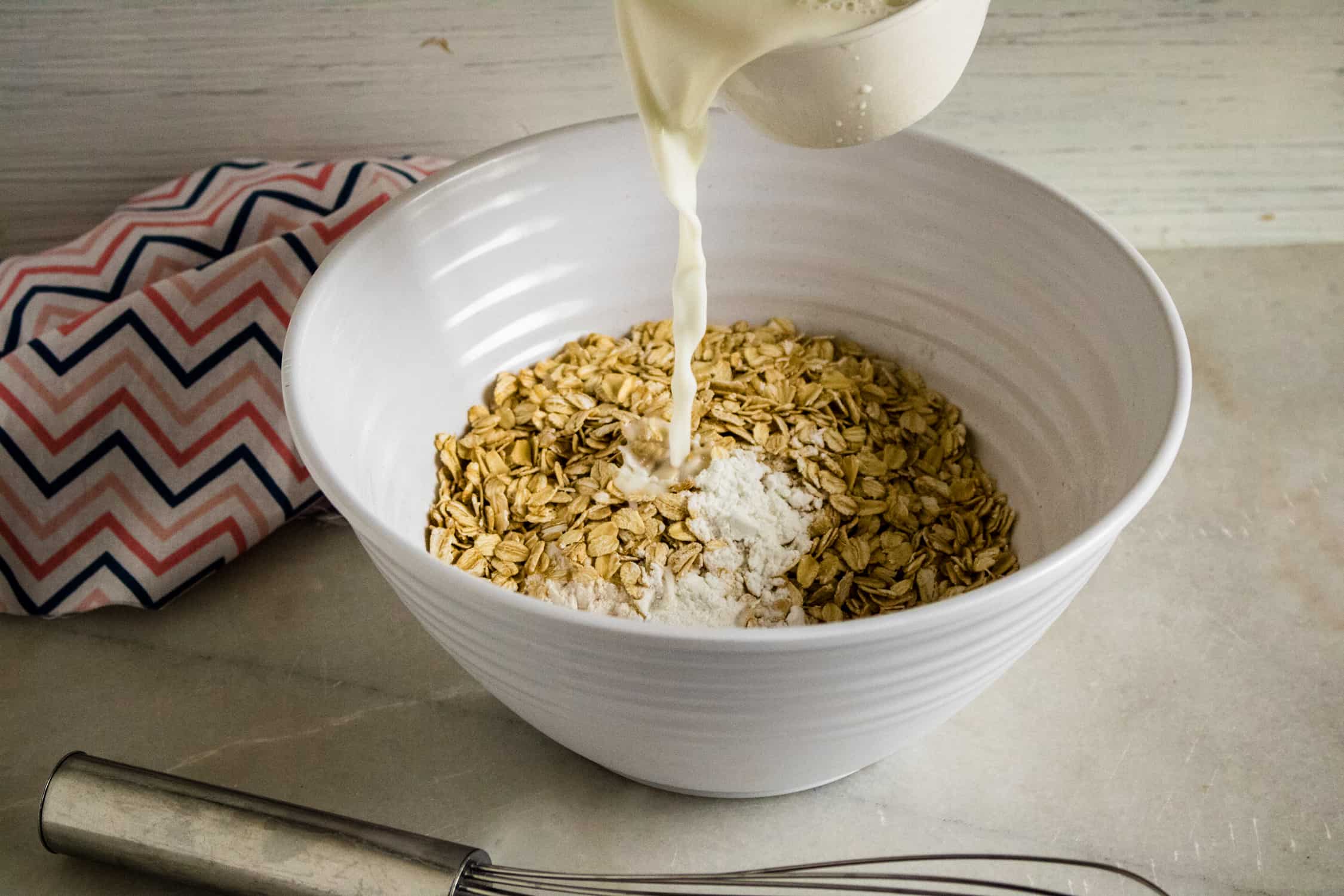 white ceramic bowl holding oats for Chocolate Chocolate Chip Oatmeal Muffins with milk being poured in from a white pitcher with silver whisk in foreground and zig zag print napkin to the left