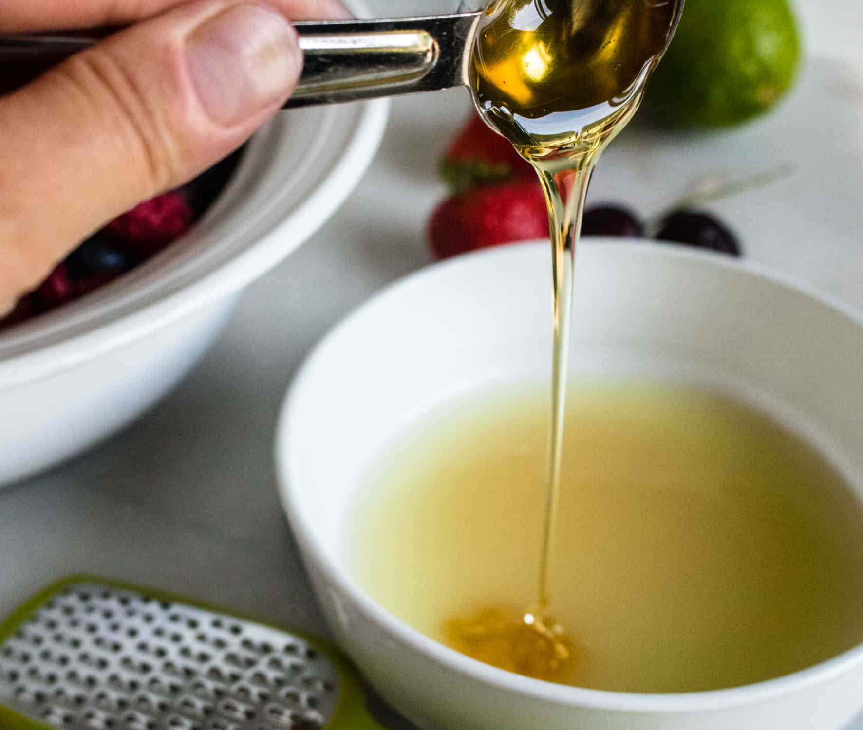 silver teaspoon of honey pouring honey into white bowl for fresh fruit salad with honey lime dressing