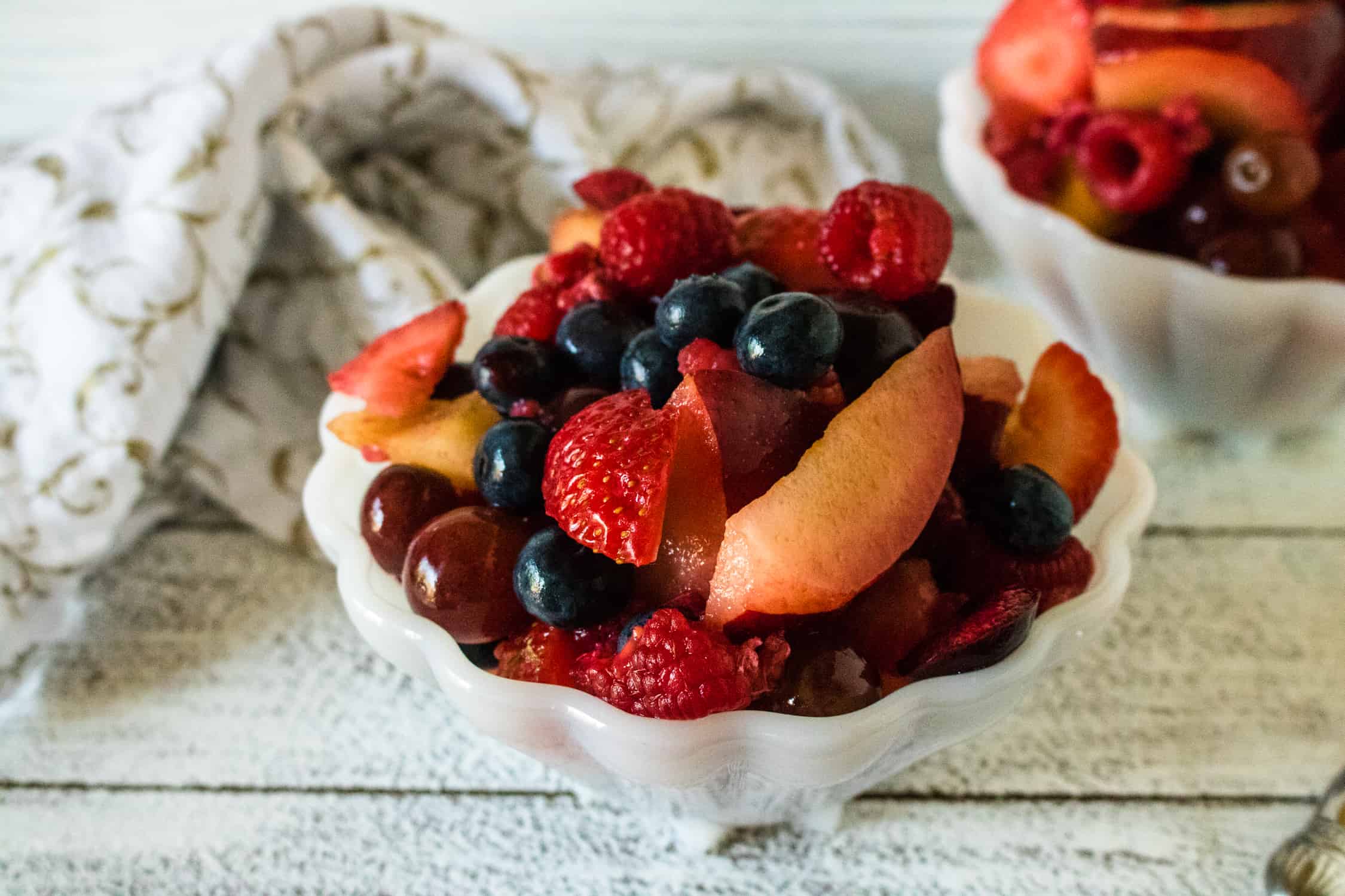 scalloped white bowl with fresh fruit salad with honey lime dressing in it with another identical bowl on background along with white and tan dishtowel