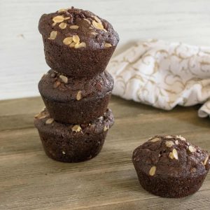 three stacked chocolate chocolate chip oatmeal muffins next to one muffin on a wood cutting board in front of a white print kitchen towel