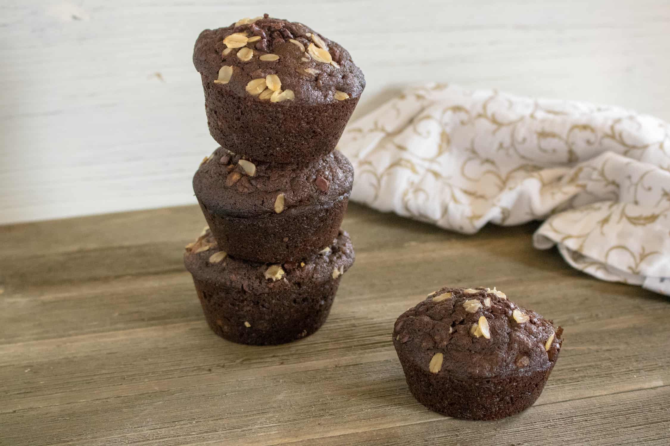3 Chocolate Chocolate Chip Oatmeal Muffins stacked on top of each other with another muffin to the right sitting on a wooden table with a white and tan dishtowel in background