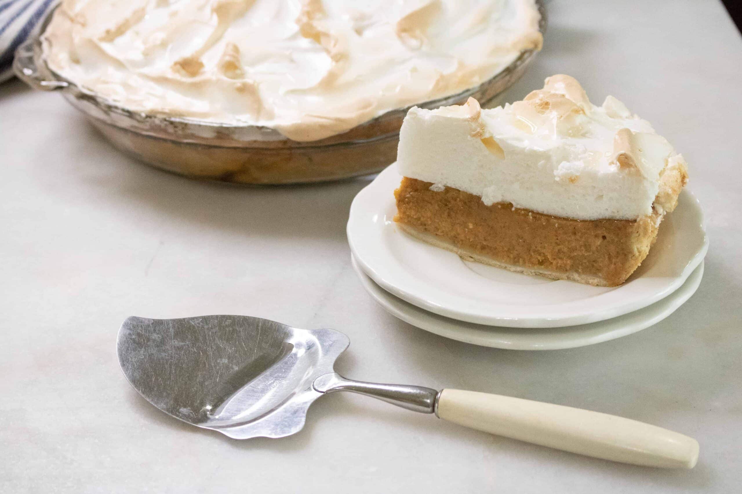 Slice of pumpkin meringue pie on stacked white plates next to a vintage pie server, with the whole pie behind it