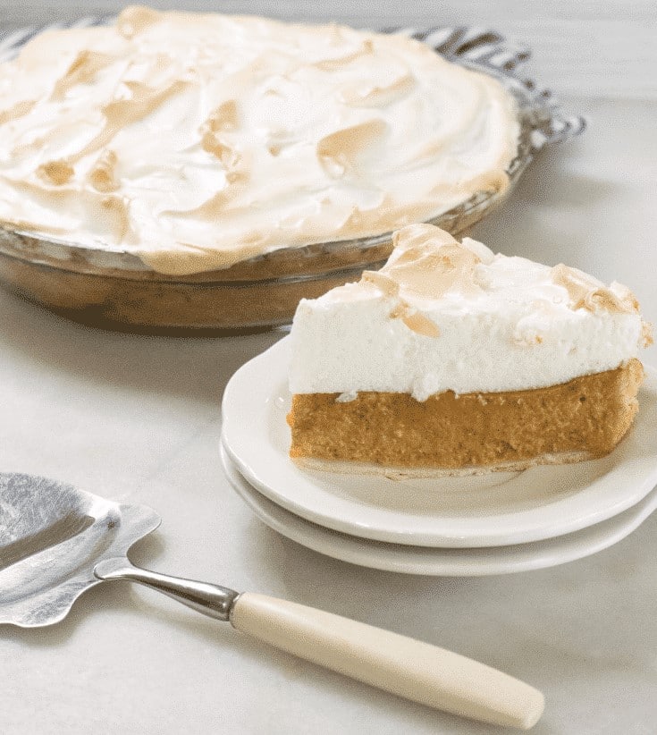 Slice of pumpkin meringue pie on a white plate in front of the whole pie, with a pie server in the foreground