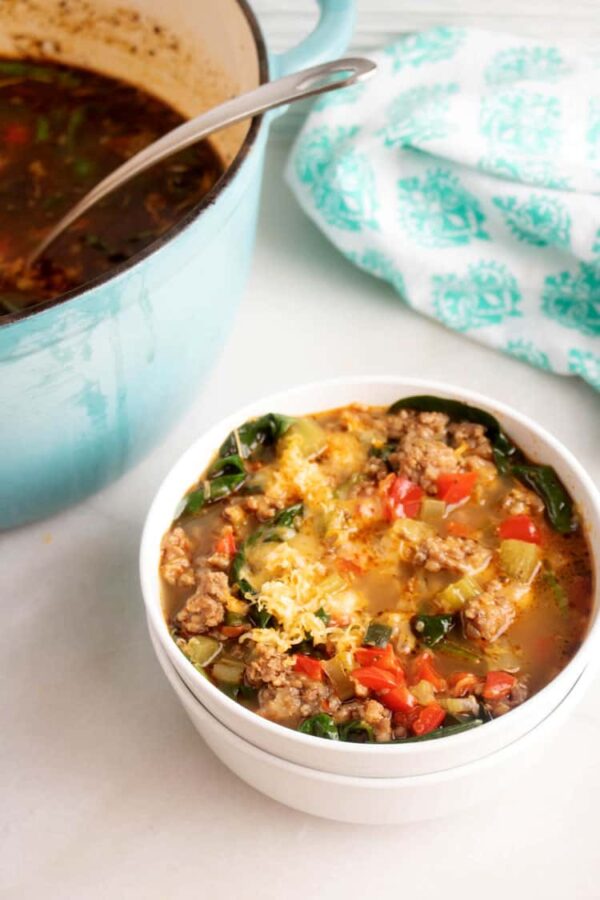Bowl of cheesy keto sausage and pepper soup next to a teal Dutch oven