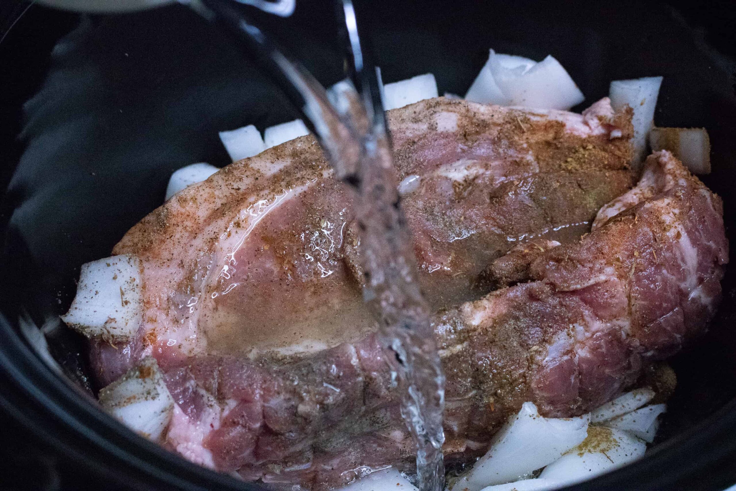 Water being poured into slow cooker on top of pork roast for Slow Cooker Pulled Pork with chopped onions and seasonings on top