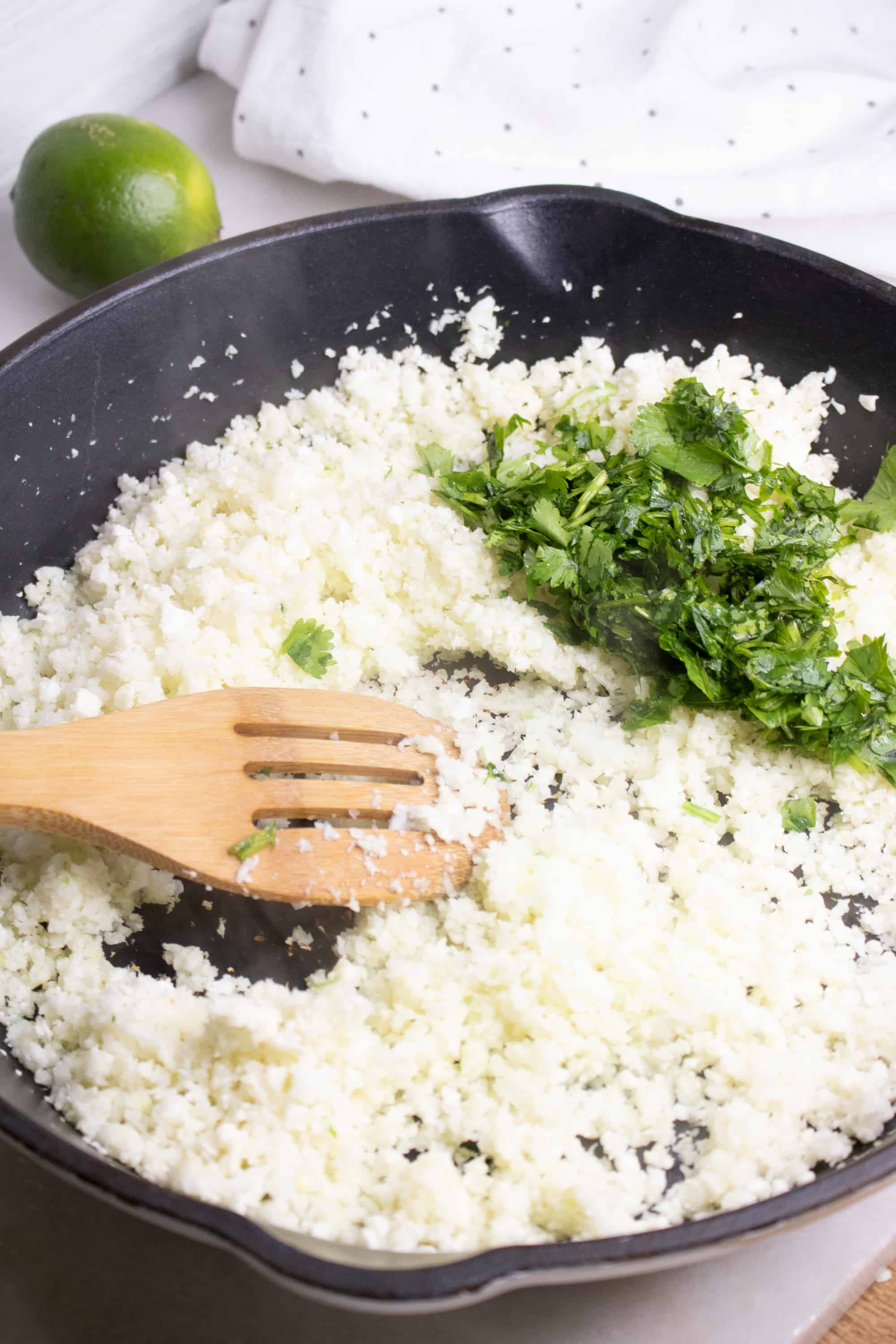 Riced cauliflower and cilantro in a gray skillet being stirred by wooden fork.