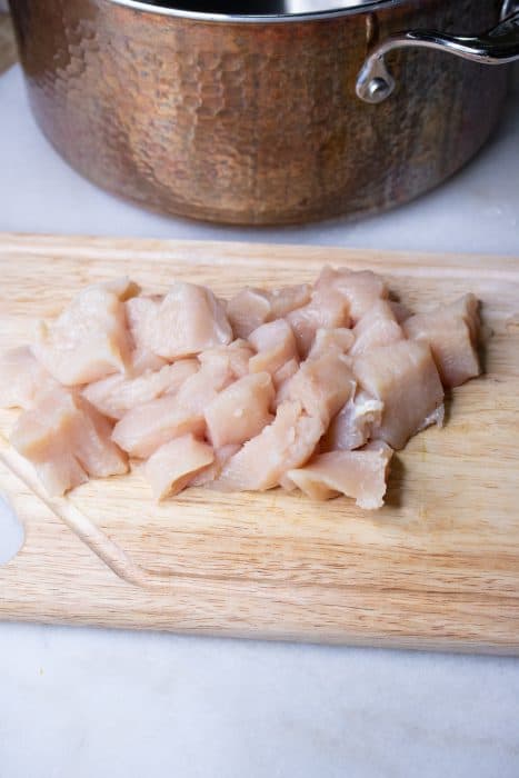 Raw chicken breast cut into cubes on a wooden cutting board with a copper pot behind it