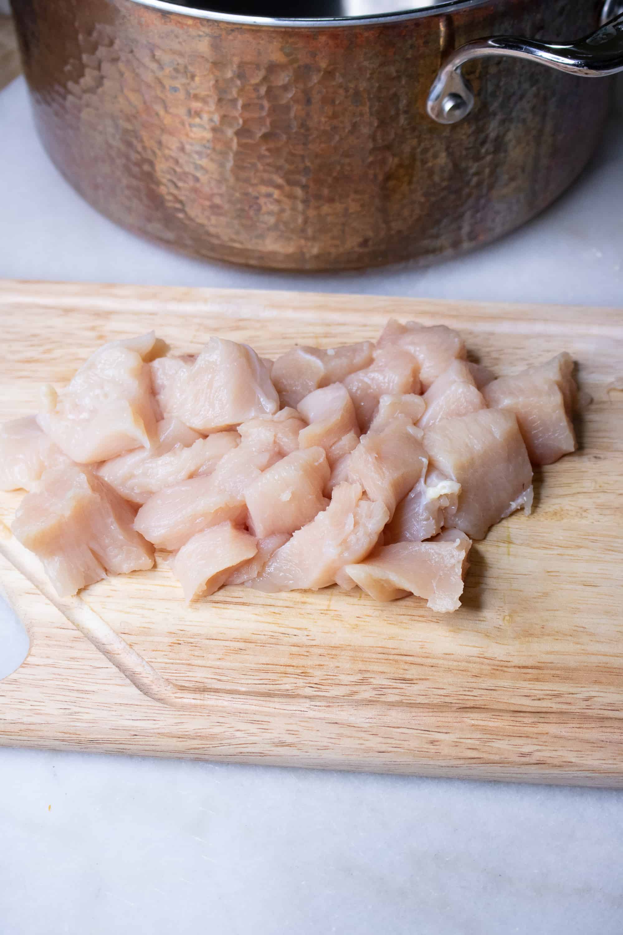 Diced raw chicken for Curry Chicken and Cauliflower Rice soup on a wooden cutting board with copper pot in background