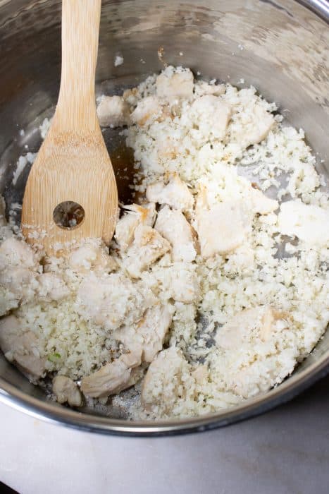 Browned chicken cubes and cauliflower rice being stirred with a wooden spoon in a stock pot