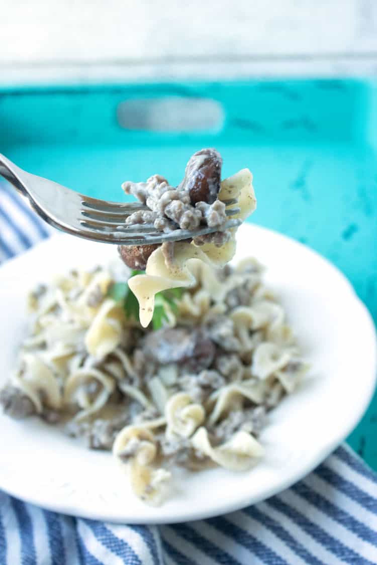 a fork full of beef stroganoff with a plate of stroganoff in the background on a white plate that is on top of a blue and white striped towel on a teal tray.