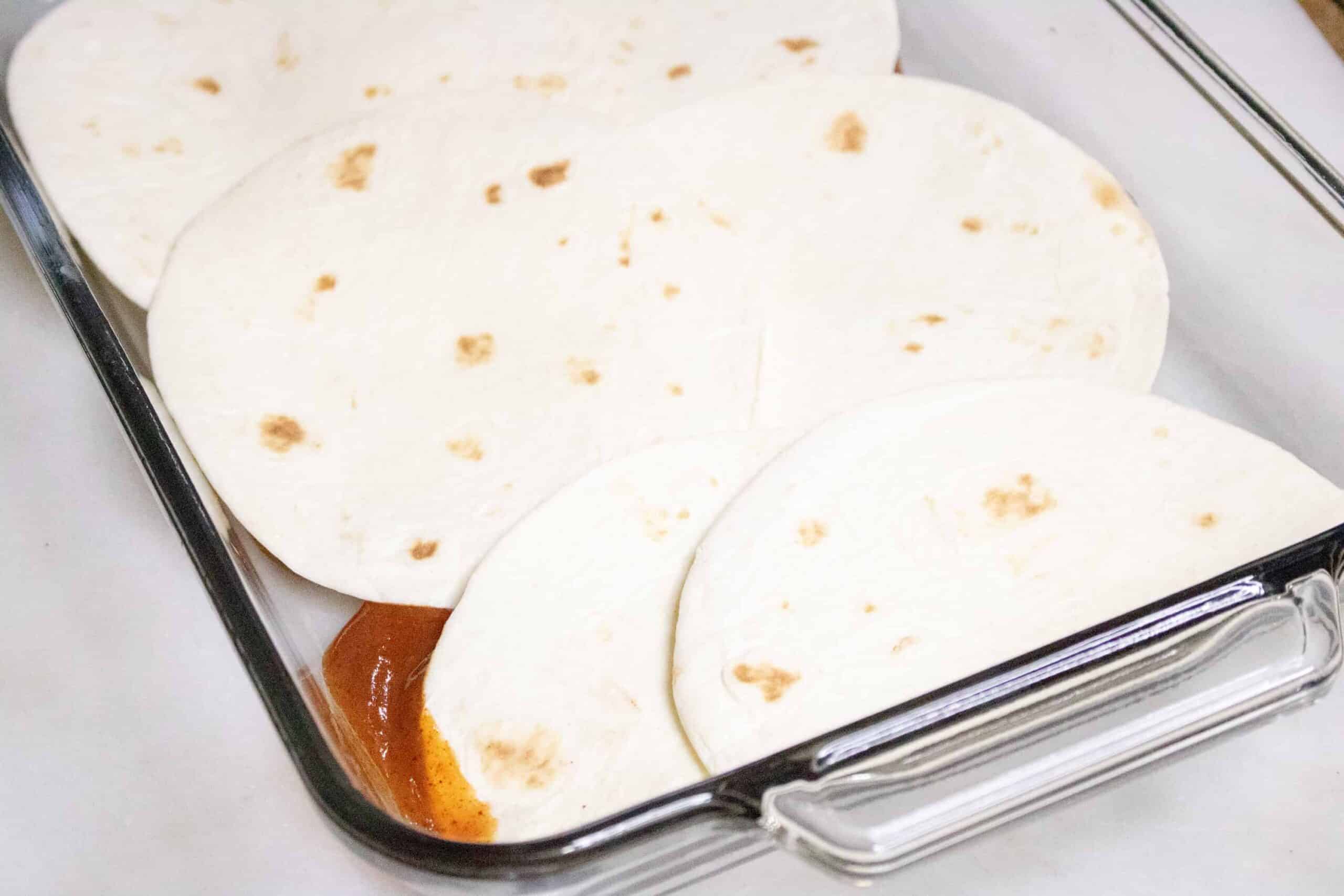 Flour tortillas for Pork Enchilada Casserole laid on top of enchilada sauce in 13 x 9 pan.