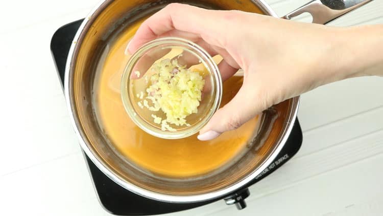 hand holding small glass bowl of minced garlic over saucepan with sauce in it