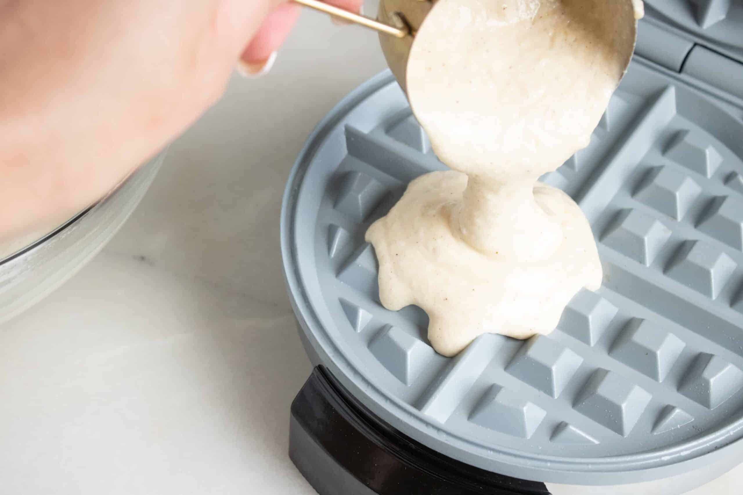 pouring a measuring cup of waffle batter into waffle iron