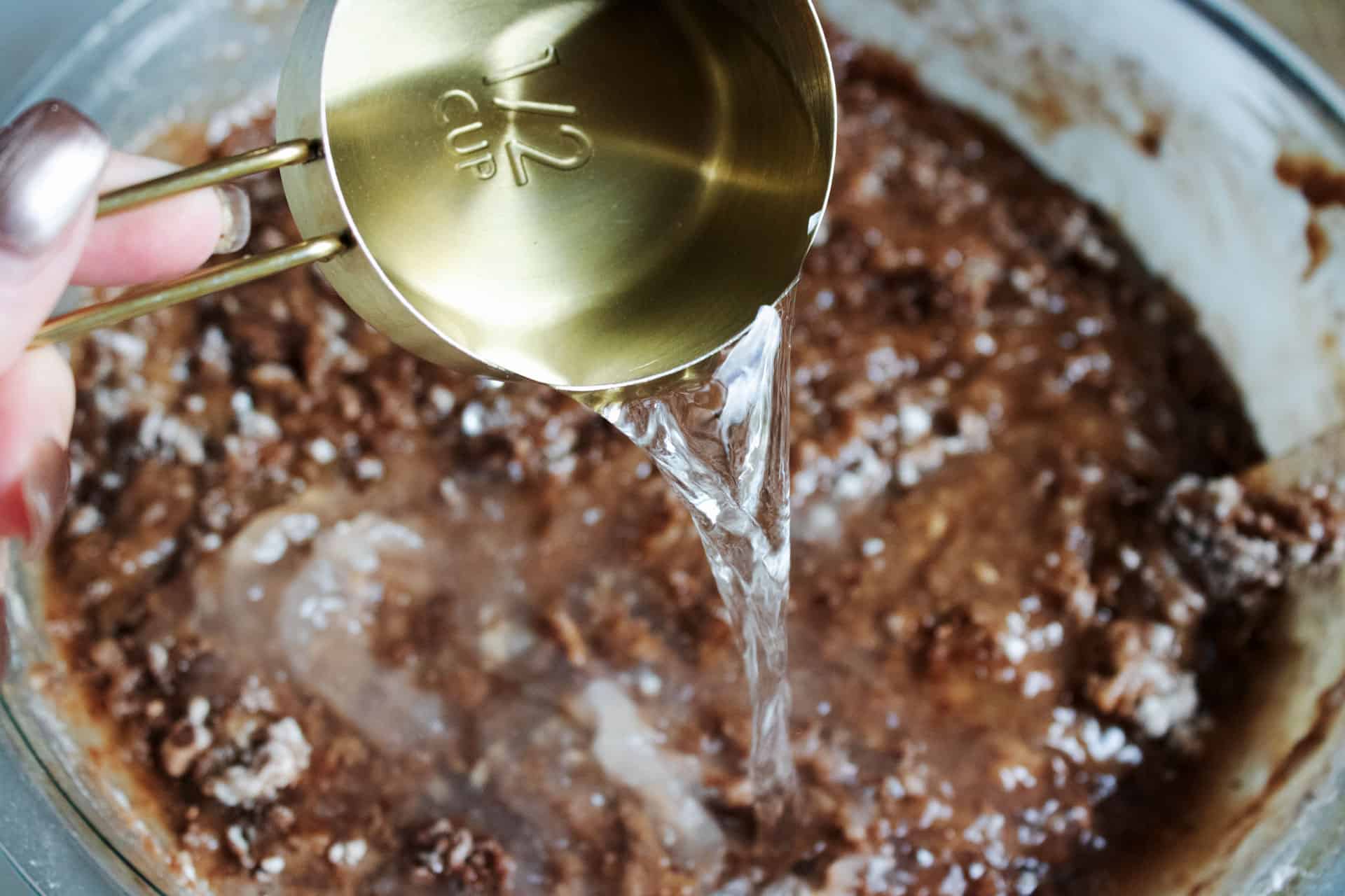 Hot water being poured into chocolate cake batter in a glass mixing bowl