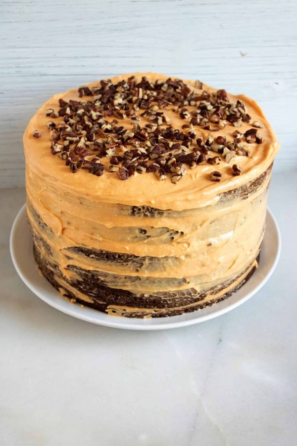 Layered chocolate cake with pumpkin cream cheese frosting topped with chopped pecans on a white plate