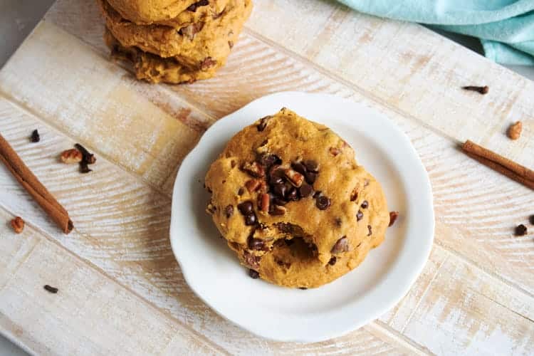 Overhead view of a pumpkin chocolate chip cookie with a bite missing on a white plate beside cinnamon sticks