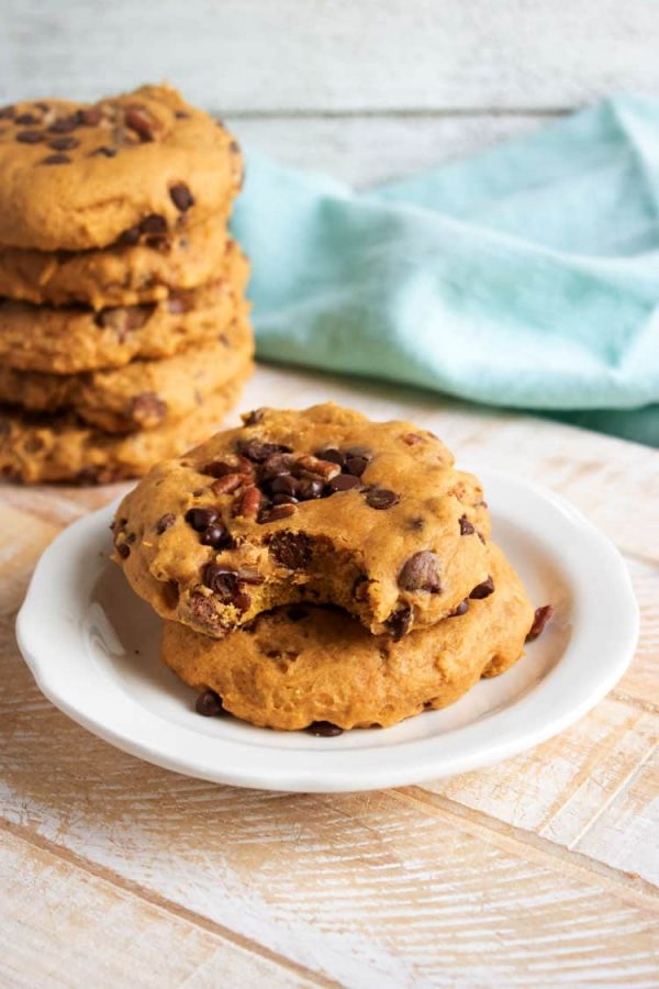 Two pumpkin chocolate chip cookies with pecans on a white plate, the top cookie bitten, with a stack of cookies behind