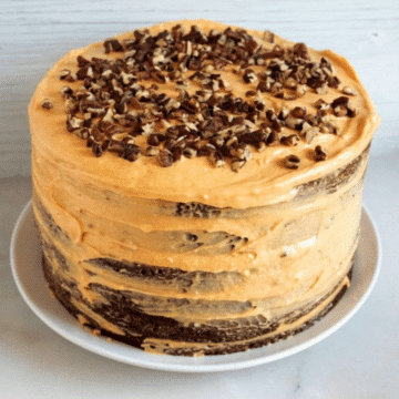 Layered chocolate cake with pumpkin cream cheese frosting topped with chopped pecans on a white plate