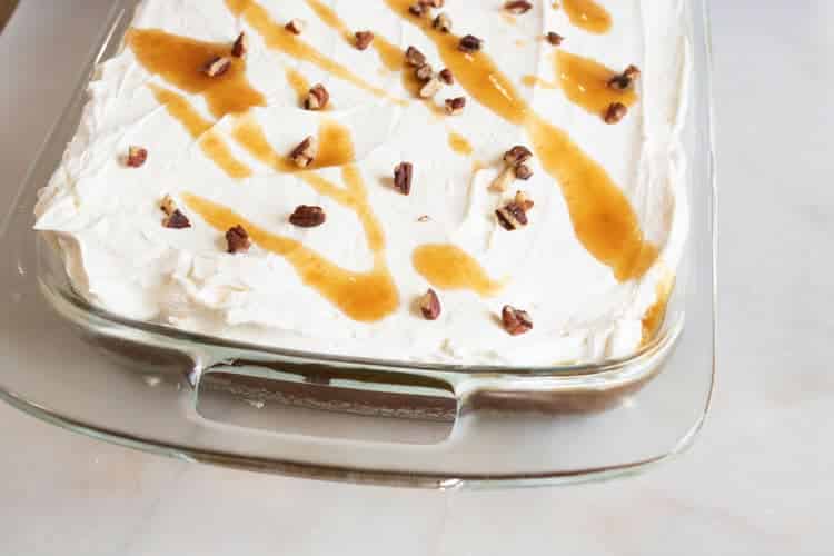 Whole pumpkin poke cake in a glass baking dish with cream cheese topping, caramel drizzle, and chopped pecans