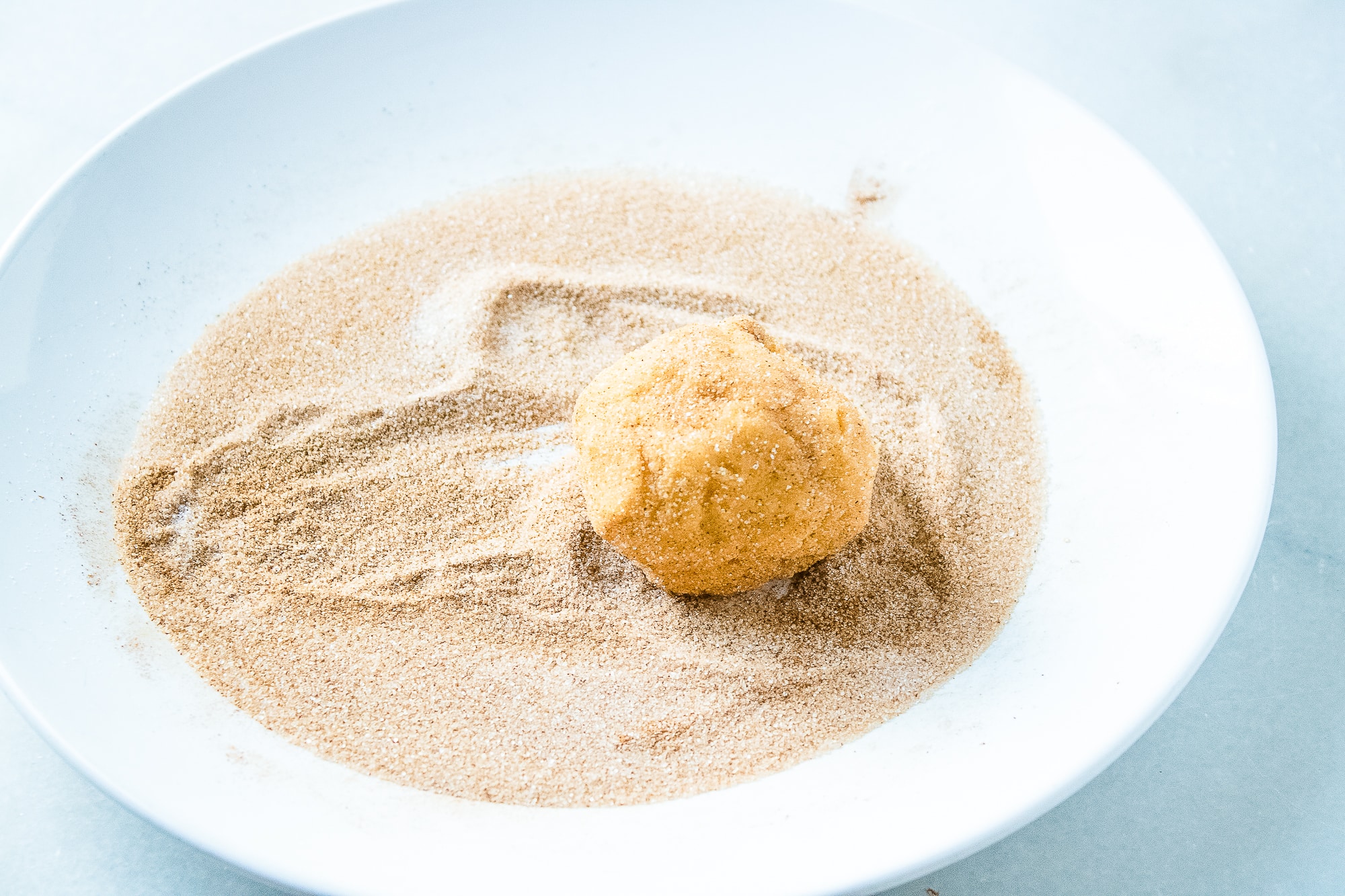 Rolling a ball of pumpkin snickerdoodle dough in cinnamon sugar on a white plate