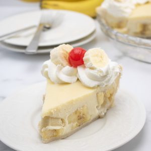 slice of banana cream pie on white plate with whipped cream, sliced bananas and a cherry on top