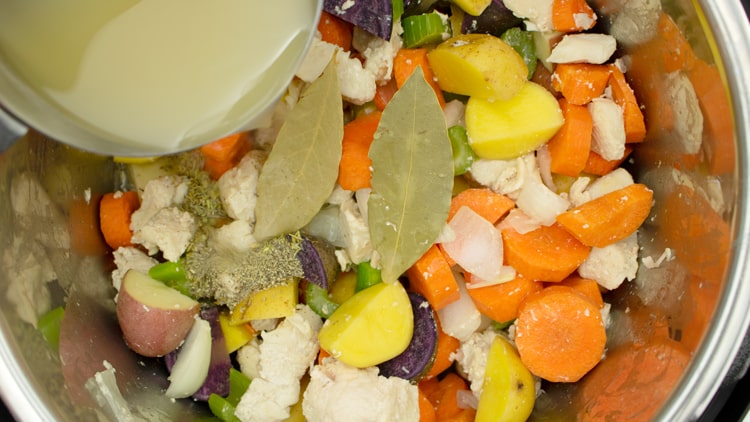 Pouring chicken stock over chicken, carrots, potatoes, bay leaves and herbs in the slow cooker