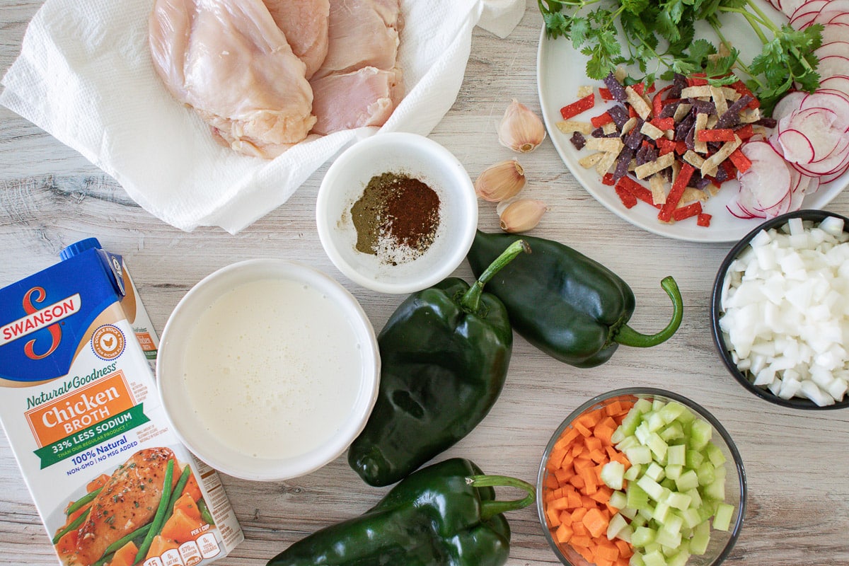 Ingredients for chicken poblano soup: raw chicken breasts, poblano peppers, diced onion, carrots and celery, chicken broth, heavy cream and spices