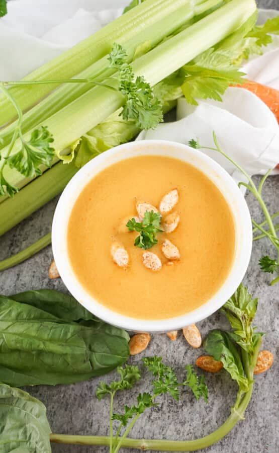 Bowl of copycat Panera autumn squash soup garnished with pumpkin seeds and parsley, surrounded by celery and carrots